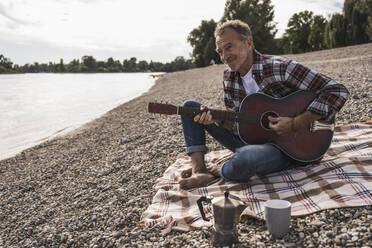 Smiling senior man playing guitar sitting at beach - UUF30842