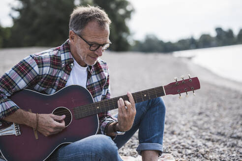 Senior man plucking guitar sitting at beach on weekend - UUF30843