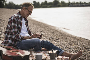 Senior man using smart phone sitting by sea at beach - UUF30844