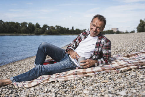 Smiling senior man with coffee cup sitting on blanket at beach - UUF30850