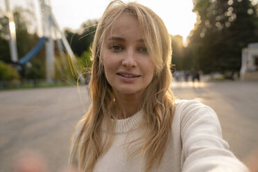 Smiling blond woman taking selfie in amusement park at sunset - VPIF08986