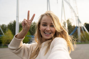 Happy blond woman taking selfie and gesturing peace sign in amusement park at sunset - VPIF08987