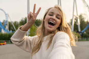 Cheerful blond woman taking selfie and gesturing peace sign in amusement park at sunset - VPIF08988