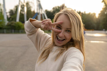 Happy woman taking selfie and gesturing peace sign in amusement park at sunset - VPIF08989