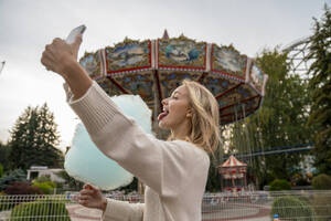 Happy woman licking cotton candy and taking selfie at amusement park - VPIF08990