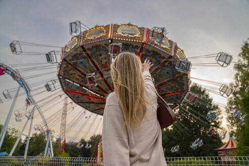 Blond woman reaching towards carousel in amusement park at sunset - VPIF08991