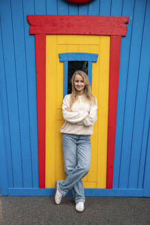 Happy young woman leaning with arms crossed on yellow wooden door - VPIF08993