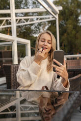 Blond woman looking at smart phone and applying lipstick in cafe - VPIF09004