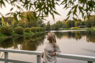 Happy woman leaning on railing in front of lake at sunset - VPIF09010