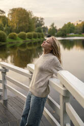 Young woman with eyes closed leaning on railing in front of lake at sunset - VPIF09012