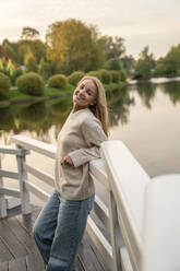 Happy young blond woman leaning on railing in front of lake at sunset - VPIF09013