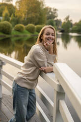 Happy blond woman leaning on railing in front of lake at sunset - VPIF09014