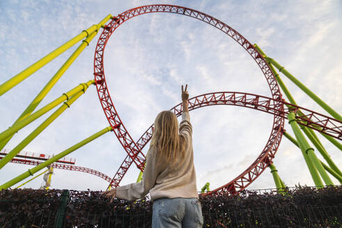 Young woman gesturing peace sign in front of rollercoaster - VPIF09019