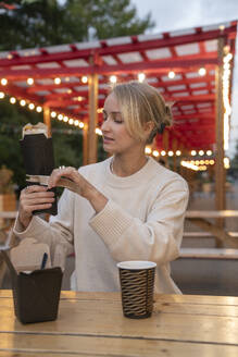 Blond young woman unwrapping food at table in amusement Park - VPIF09028