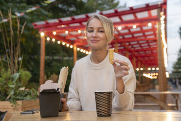 Smiling blond woman eating food at table in amusement Park - VPIF09029