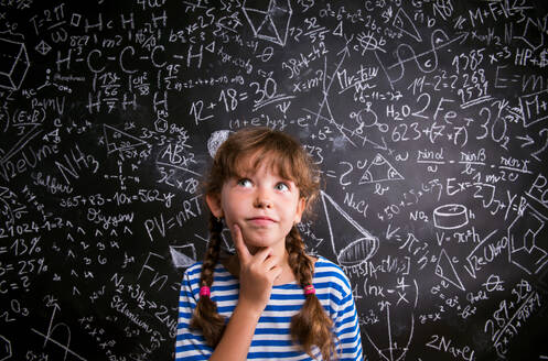Thinking girl in blue striped t-shirt with two braids with finger on her cheek against big blackboard with mathematical symbols and formulas - HPIF35385