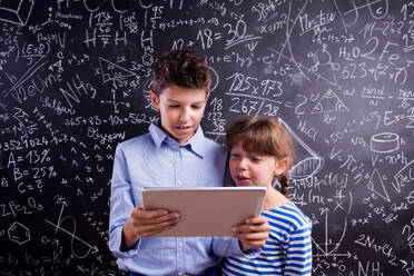 Cute boy and girl with tablet at school in front of a big blackboard. Studio shot on black background. - HPIF35415