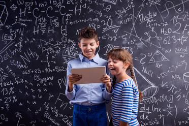 Cute boy and girl with tablet at school in front of a big blackboard. Studio shot on black background. - HPIF35416