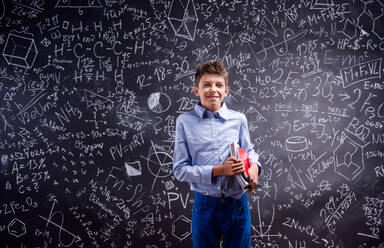 Junge im blauen Hemd hält verschiedene Notizbücher an eine große Tafel mit mathematischen Symbolen und Formeln - HPIF35425