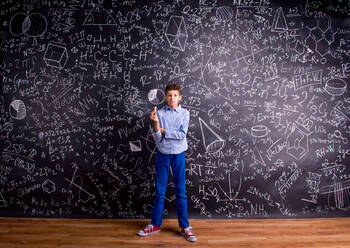 Junge in blauem T-Shirt, der ein Stück Kreide hält, vor einer großen Tafel mit mathematischen Symbolen und Formeln - HPIF35441