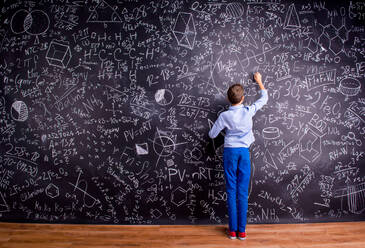 Junge in blauem T-Shirt schreibt mit Kreide auf große Tafel mit mathematischen Symbolen und Formeln, Rückansicht - HPIF35442