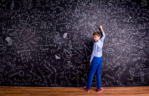 Junge in blauem T-Shirt schreibt mit Kreide auf große Tafel mit mathematischen Symbolen und Formeln, Rückansicht - HPIF35444