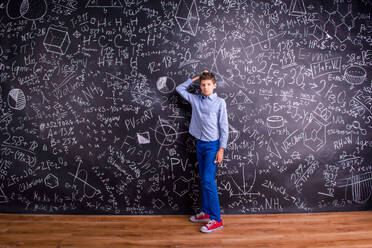 Junge in blauem T-Shirt, kratzt sich am Kopf, vor einer großen Tafel mit mathematischen Symbolen und Formeln - HPIF35445