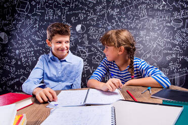 Young school boy and girl sitting at the desk, doing their homework against big blackboard with formulas and mathematical symbols - HPIF35478