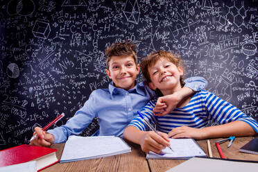 Junge Schule Junge und Mädchen sitzen am Schreibtisch, machen ihre Hausaufgaben gegen große Tafel mit Formeln und mathematische Symbole - HPIF35479