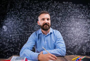 Happy hipster teacher sitting at the desk with various school supplies, against big blackboard with formulas and mathematical symbols - HPIF35483