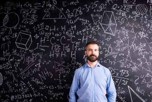 Hipster teacher standing against big blackboard with mathematical symbols and formulas. Studio shot on black background. - HPIF35505
