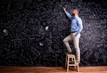 Hipster teacher writing on big blackboard with mathematical symbols and formulas, standing on step ladder. Studio shot on black background. - HPIF35506