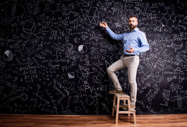 Hipster teacher writing on big blackboard with mathematical symbols and formulas, standing on step ladder. Studio shot on black background. - HPIF35507