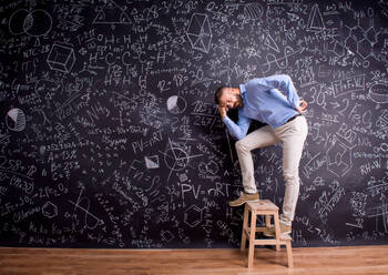 Hipster-Lehrer auf Stufenleiter stehend, vor großer Tafel mit mathematischen Symbolen und Formeln. Studioaufnahme auf schwarzem Hintergrund. - HPIF35508