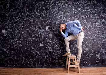 Hipster teacher standing on step ladder, against big blackboard with mathematical symbols and formulas. Studio shot on black background. - HPIF35508