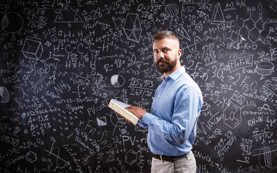 Hipster-Lehrer mit Buch vor großer Tafel mit mathematischen Symbolen und Formeln, Studioaufnahme auf schwarzem Hintergrund. - HPIF35519