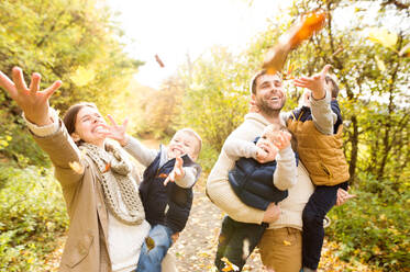Schöne junge Familie auf einem Spaziergang im bunten Herbstwald. Mutter und Vater tragen ihre drei Söhne in den Armen und werfen Blätter in die Luft. - HPIF35563
