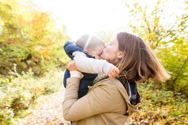 Schöne junge Mutter mit ihrem Sohn in warmer Kleidung auf einem Spaziergang draußen in der bunten Herbstnatur, hält ihn in ihren Armen, küsst ihn. - HPIF35566
