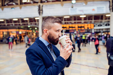 Hipster-Geschäftsmann hält einen Einwegbecher und trinkt Kaffee auf dem überfüllten Bahnhof - HPIF35773