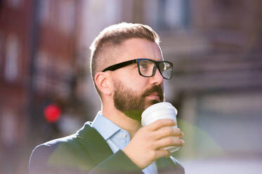 Hipster-Geschäftsmann, der einen Einwegbecher in der Hand hält und Kaffee trinkt, auf der Straße in London, mit schwarzer Brille - HPIF35837