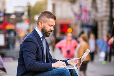 Hipster-Manager sitzt auf einer Treppe am sonnigen Piccadilly Circus, London, und arbeitet am Laptop - HPIF35856