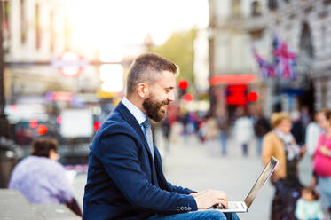 Hipster-Manager sitzt auf einer Treppe am sonnigen Piccadilly Circus, London, und arbeitet am Laptop - HPIF35859