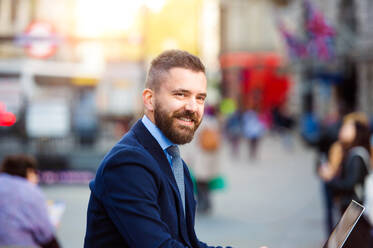 Hipster-Manager sitzt auf einer Treppe am sonnigen Piccadilly Circus, London, und arbeitet am Laptop - HPIF35861