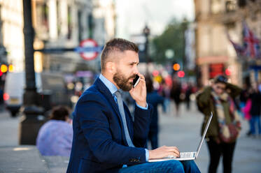 Hipster-Manager sitzt auf einer Treppe am sonnigen Piccadilly Circus, London, arbeitet am Laptop und benutzt ein Smartphone - HPIF35862