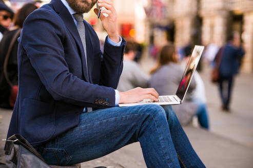 Unbekannter Manager sitzt auf einer Treppe am sonnigen Piccadilly Circus, London, arbeitet am Laptop und spricht mit einem Smartphone - HPIF35863