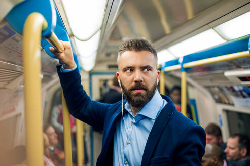 Seriöser Geschäftsmann mit Kopfhörern auf dem Weg zur Arbeit. Steht in einem U-Bahn-Waggon und hält einen Handgriff. - HPIF35880
