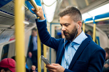 Serious businessman with headphones travelling to work. Standing inside underground wagon, holding handhandle. - HPIF35881