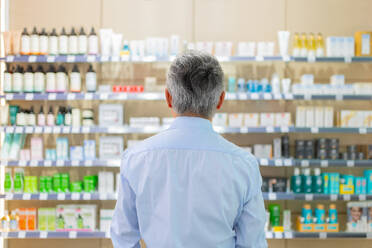 Back view of anonymous senior man with gray hair standing and choosing from assorted products on shelves in pharmaceutical store - ADSF50367