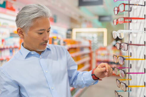 Handsome calm senior man with gray hair dressed in shirt choosing eyewear while standing near rack during shopping at optical store - ADSF50368