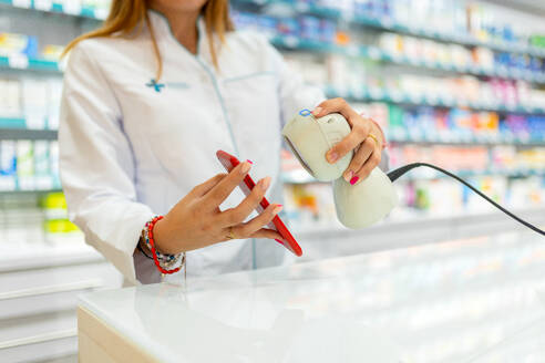 Crop hand of anonymous female cashier wearing various bracelets scanning smart phone with barcode reader in pharmacy store - ADSF50371